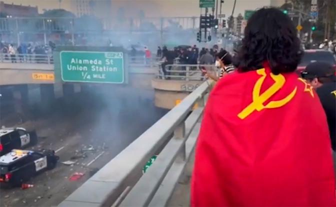 На фото: мужчина с советским флагом на спине рассматривает блокаду полицейских машин, устроенную протестующими.